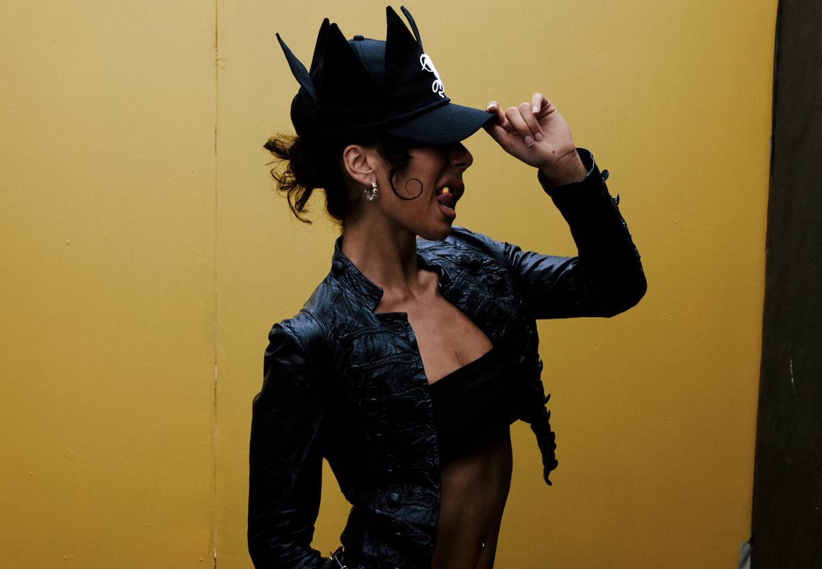 Musician Miss Kaninna posing against a bright yellow backdrop, wearing a black leather jacket and black crown-like hat.